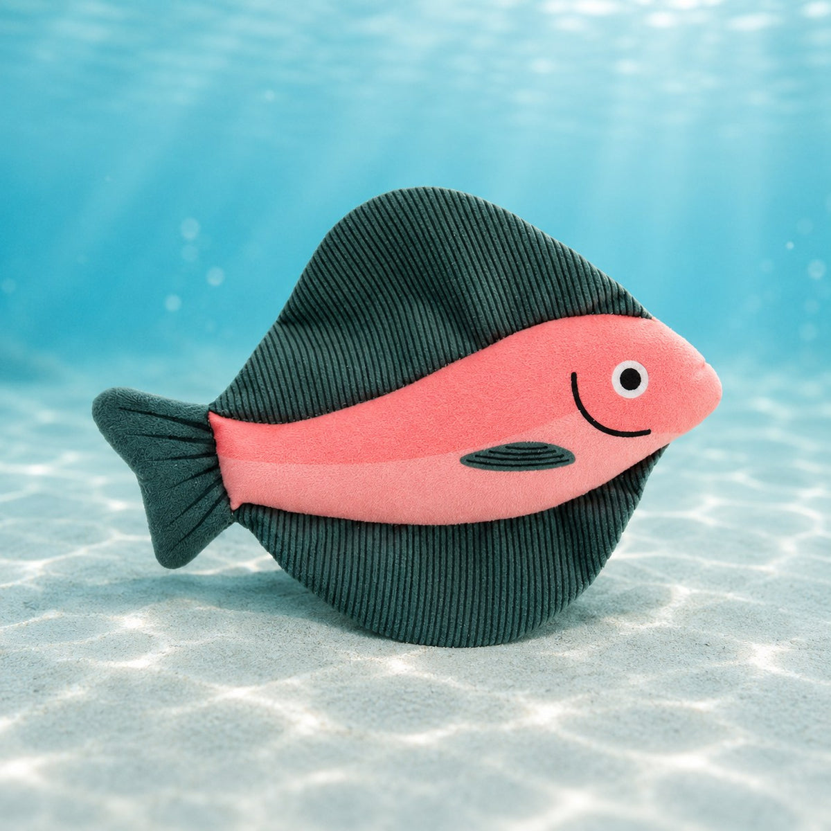 水中を背景にしたピンクの魚（ヒラメ柄）のぬいぐるみ単体カット。濃緑のコード生地の背びれと薄ピンクの体、笑顔の刺繍が見える