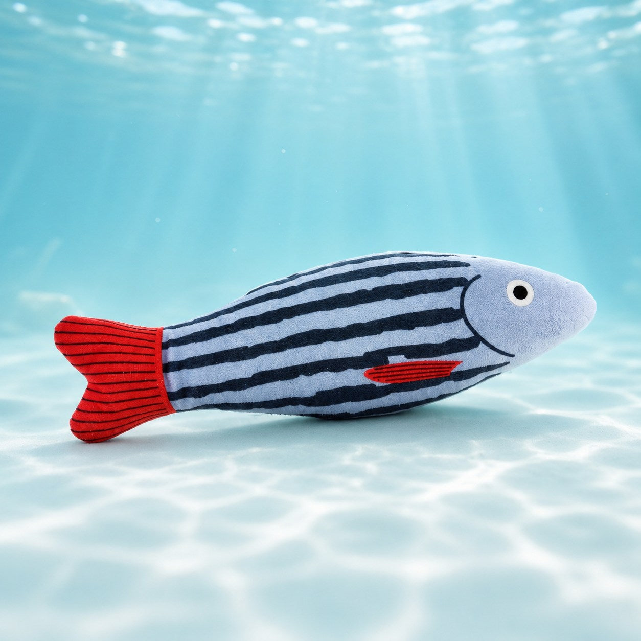 水中を背景にした縞の魚のぬいぐるみ単体カット。水色に紺の縞模様の胴体と赤いしっぽ、笑った表情のアップ