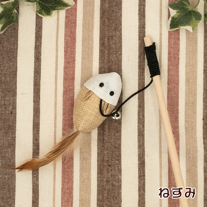 ねずみ型チャーム。白い頭部と麻布の胴体、茶色い羽根の尻尾、鈴付き。木製の柄とストライプ生地の上に配置