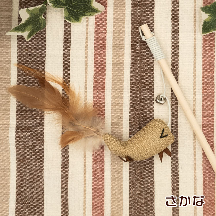 さかな型チャーム。麻布に目と口を刺繍した魚の形、茶色い羽根の尾ひれ、鈴付きの木製の柄とともにストライプ生地の上に配置