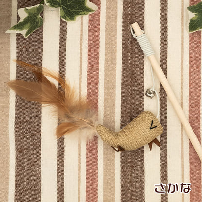 さかな型チャーム。麻布に目と口を刺繍した魚の形、茶色い羽根の尾ひれ、鈴付きの木製の柄とともにストライプ生地の上に配置