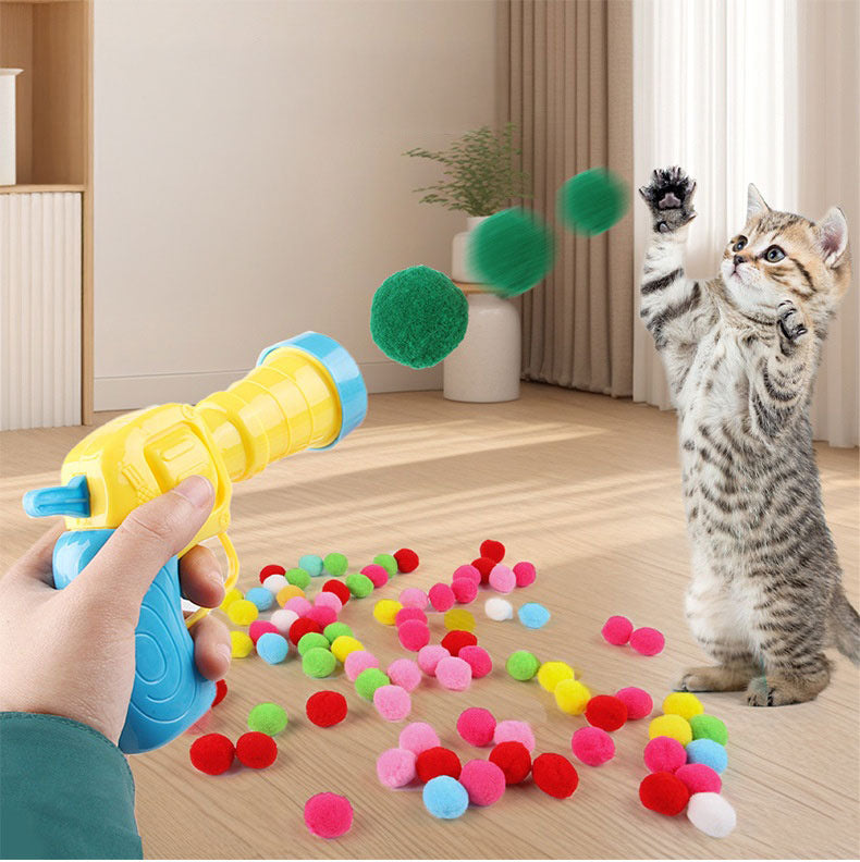 室内でガンから緑のポンポンボールが連続発射され、後ろ足で立った子猫が前足を伸ばして追いかける様子。床にはカラフルなボールが散らばる