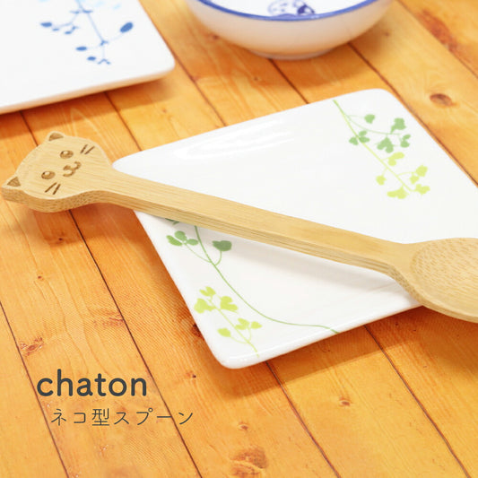 木目の食卓に置かれたchaton ネコ型スプーン。柄の先端が猫の顔になっており、緑の葉柄の白い角皿に立てかけてある
