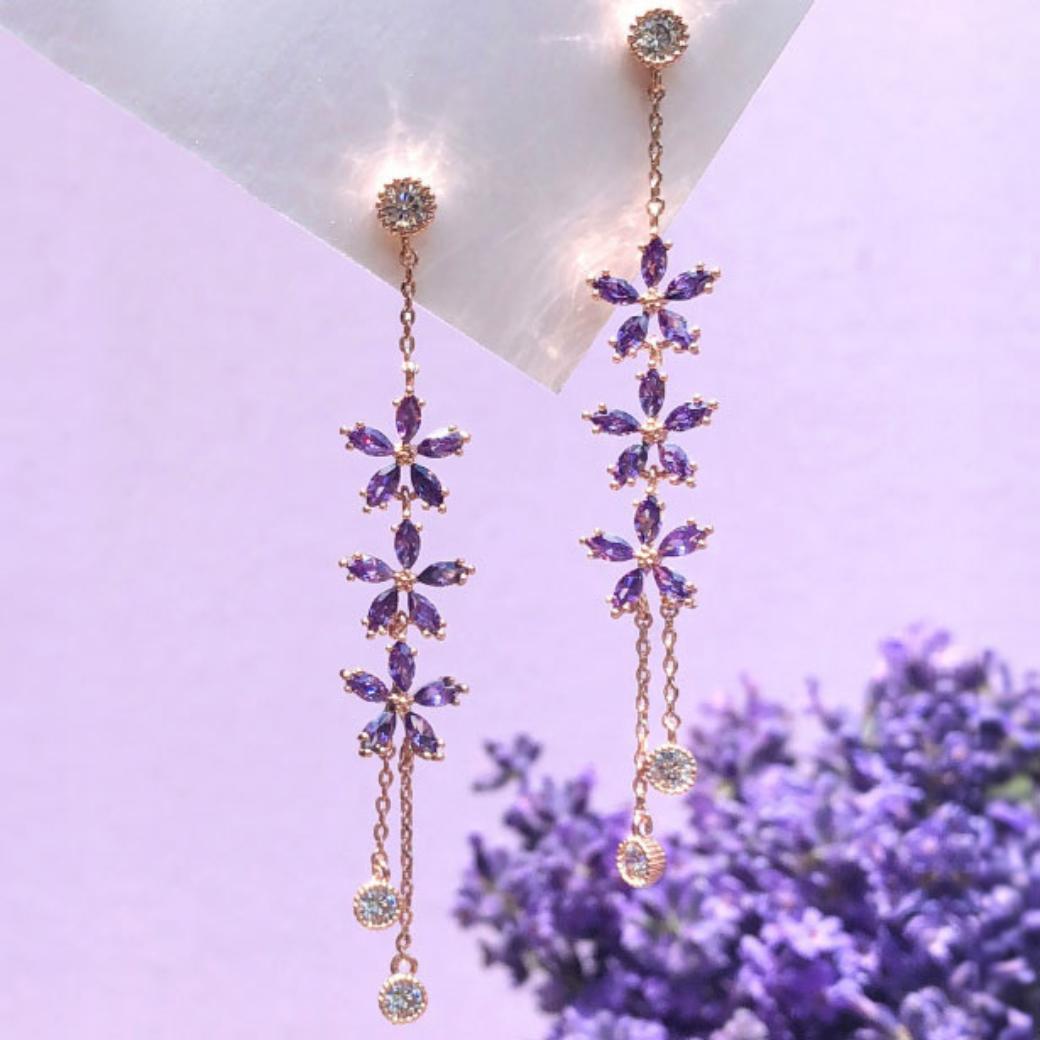 紫の花が3つ連なるロングピアス2本を、ラベンダーの花と半透明の紙を背景に並べた商品写真