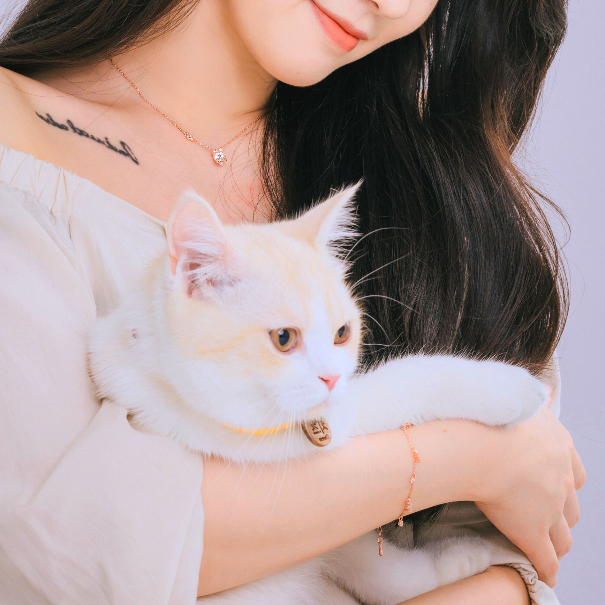 白猫を抱っこするモデルの腕にローズゴールドのブレスレット、猫の首輪にはネームプレートが揺れる