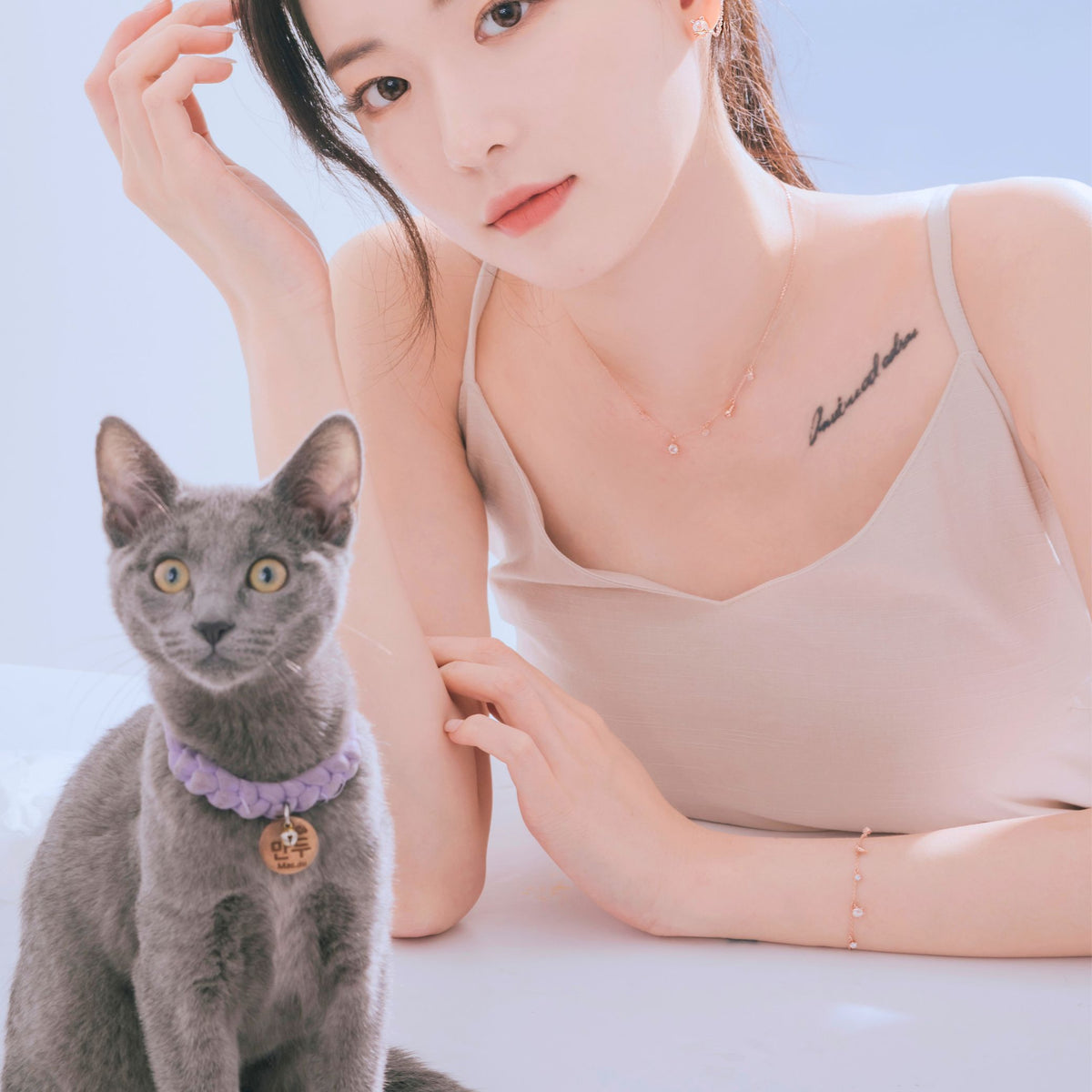 グレーの猫と並ぶモデルの腕にブレスレット、猫の首には紫の編み込みカラーとネームタグ