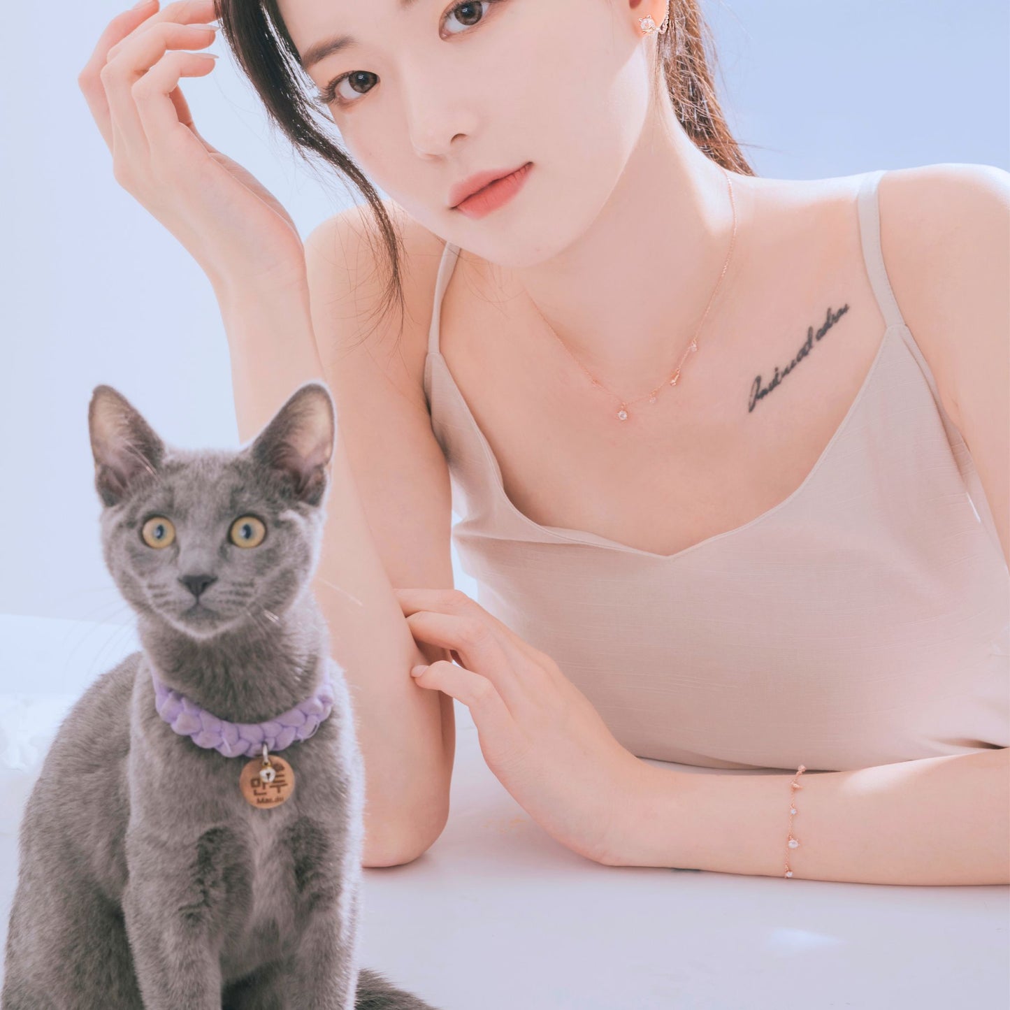 グレーの猫と並ぶモデルの腕にブレスレット、猫の首には紫の編み込みカラーとネームタグ