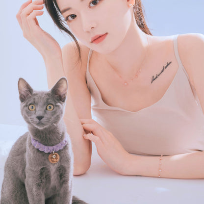 グレーの猫と並ぶモデルの腕にブレスレット、猫の首には紫の編み込みカラーとネームタグ
