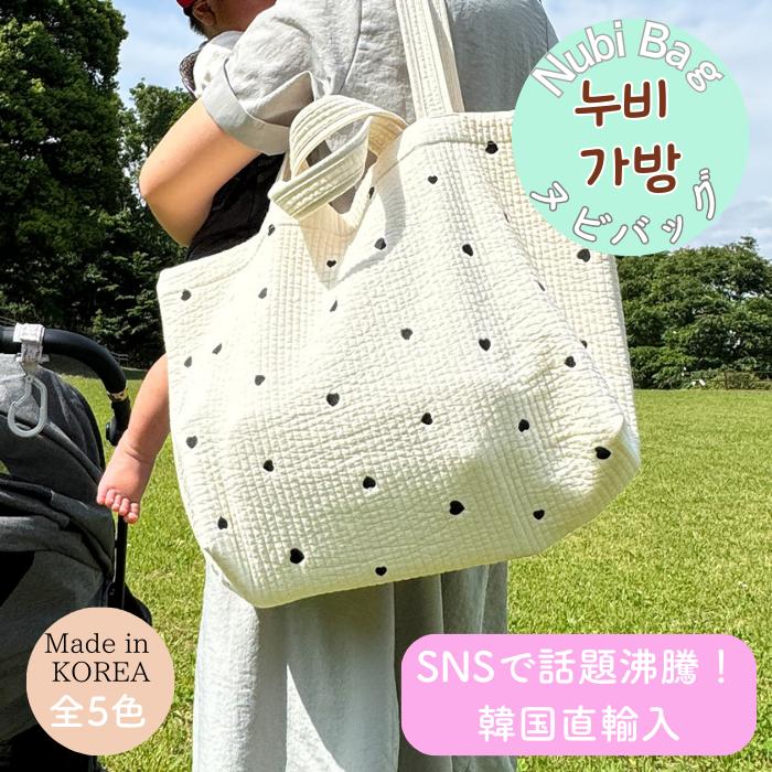 ベビーカーの脇で赤ちゃんを抱っこしながらヌビバッグをハート柄で肩に掛ける親。公園の芝生を背景に大容量トートを持ち歩く様子